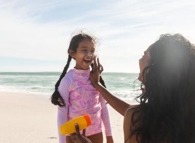 Mom and school-aged daughter apply sunscreen on beach.